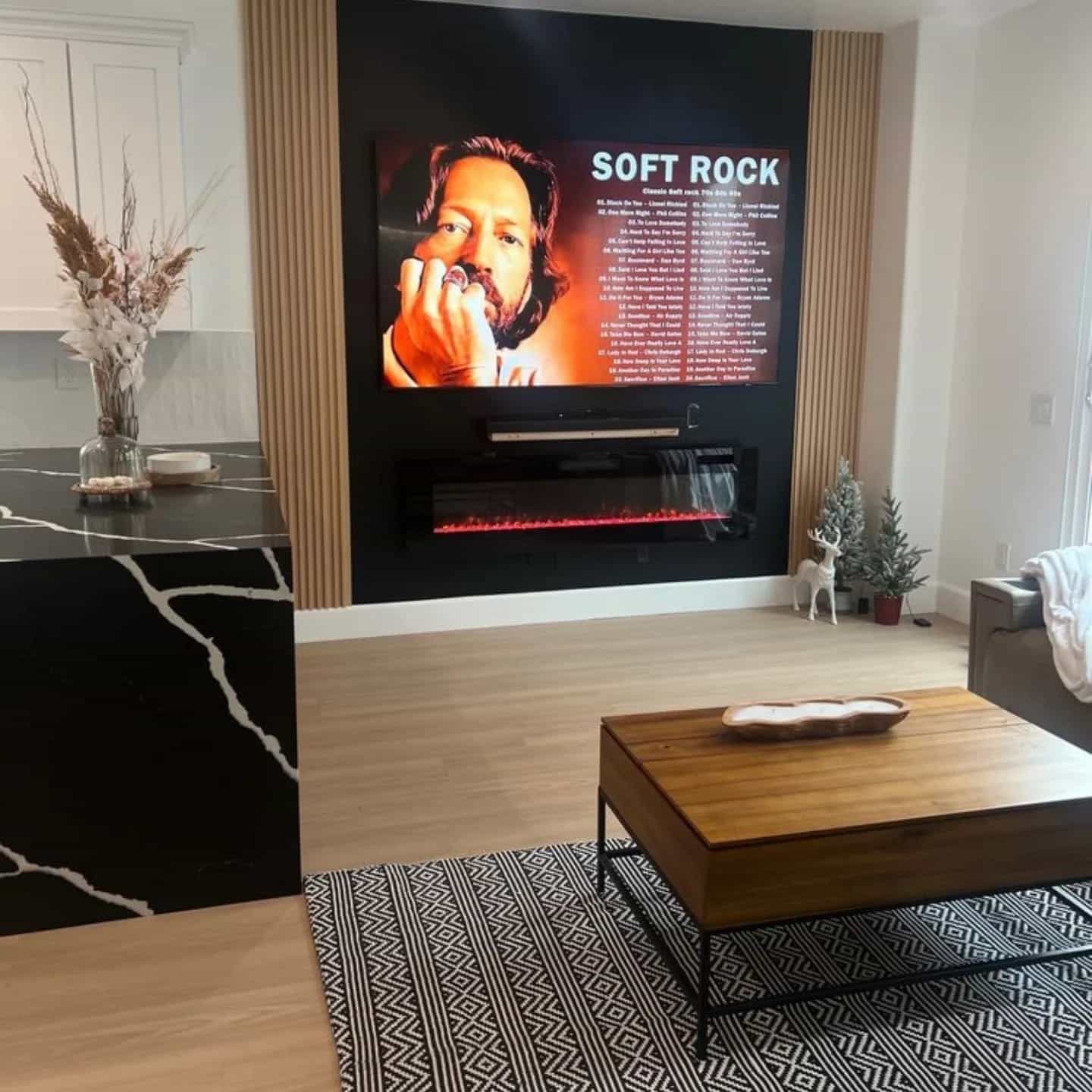 A modern living room features a large wall-mounted television displaying a "Soft Rock" music playlist with an artist's portrait. The space includes a wooden coffee table with a decorative bowl, a black accent chair, light wood flooring with geometric patterned rugs, and warm beige curtains framing the entertainment wall. Decorative elements such as potted plants, a white decorative deer figurine, and a vase with dried branches complete the contemporary interior design.