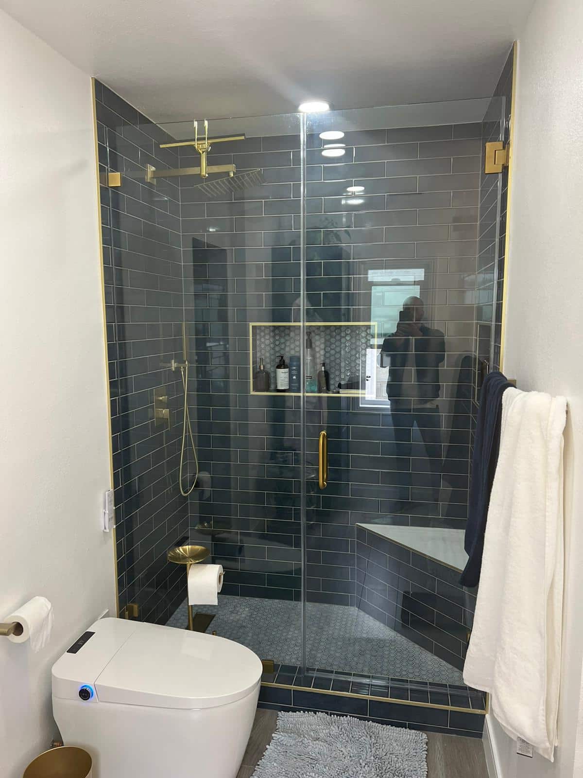 A modern bathroom remodel featuring a spacious walk-in shower with dark gray subway tile walls, brass hardware, and a frameless glass enclosure with a bench seat. The sleek white bidet toilet, heated towel rack, and coordinating brass fixtures complement the contemporary design, while soft gray tile flooring and bright LED lighting complete the upscale aesthetic.