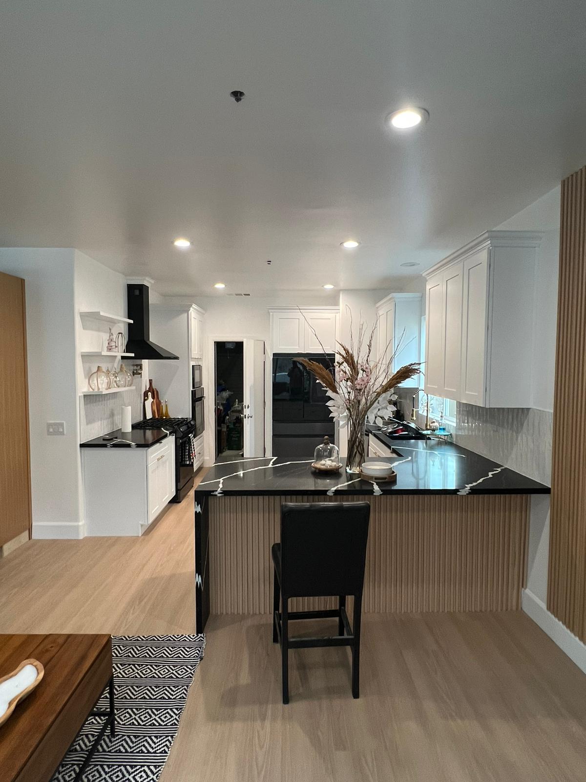 A modern kitchen remodel featuring white cabinetry, black countertops, and a large island with seating, accented by warm wood flooring and stainless steel appliances including a black range hood. The space showcases recessed lighting, a geometric patterned runner rug, and decorative elements like a vase with dried florals. Floor-to-ceiling white cabinetry and a black interior door frame the galley-style layout, creating a contemporary and functional kitchen design.