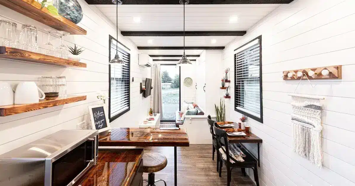 A modern farmhouse kitchen features white shiplap walls, rich wooden countertops and shelving, and black-framed windows overlooking outdoor scenery. The galley-style layout includes pendant lighting, stainless steel appliances, decorative wooden shelves displaying dishware and plants, and woven textile accents. Dark wood flooring and natural materials create a warm, contemporary aesthetic ideal for a kitchen remodel.