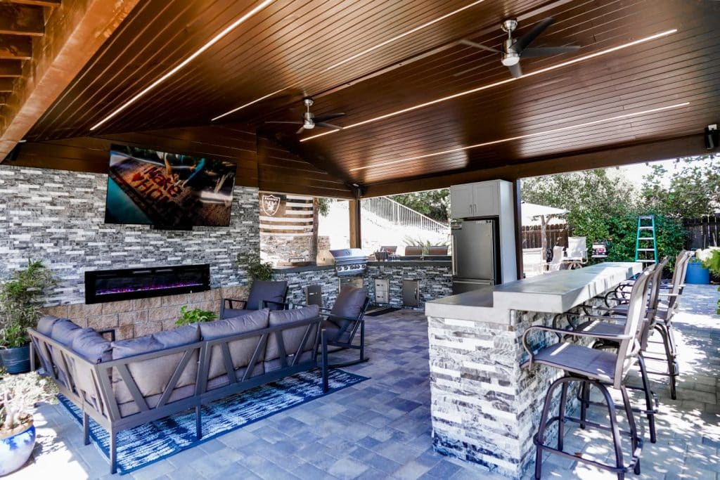 A modern covered outdoor living space featuring a lounge area with purple cushioned seating, a linear electric fireplace on a stacked stone accent wall, and a flat-screen TV mounted above. The adjacent kitchen and dining zone includes a built-in grill, stainless steel appliances, and bar seating with views to a backyard patio, all unified by a wood-slat ceiling, LED lighting, and decorative tile flooring.