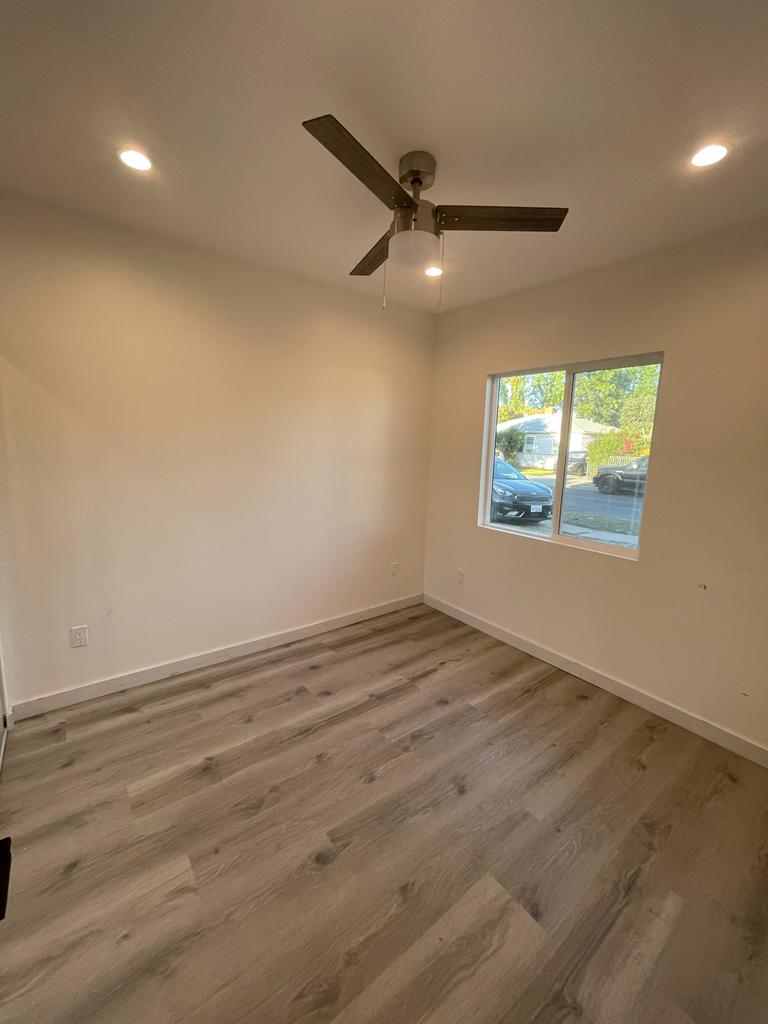 An empty bedroom interior with light wood-look laminate flooring, cream-colored walls, and a ceiling fan with recessed lighting. A single window with blue trim lets in natural light, suggesting a newly remodeled or staged residential space ready for occupancy.