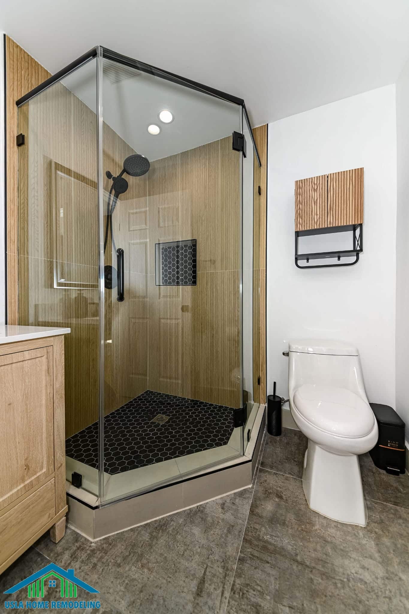 A modern bathroom remodel featuring a spacious walk-in shower with a glass enclosure, black hexagonal floor tiles, and wood-textured walls. The bathroom includes a white toilet, wooden vanity with sink on the left, and a wood and metal wall shelf above the toilet. Gray wood-look flooring and white walls complete the contemporary design aesthetic.