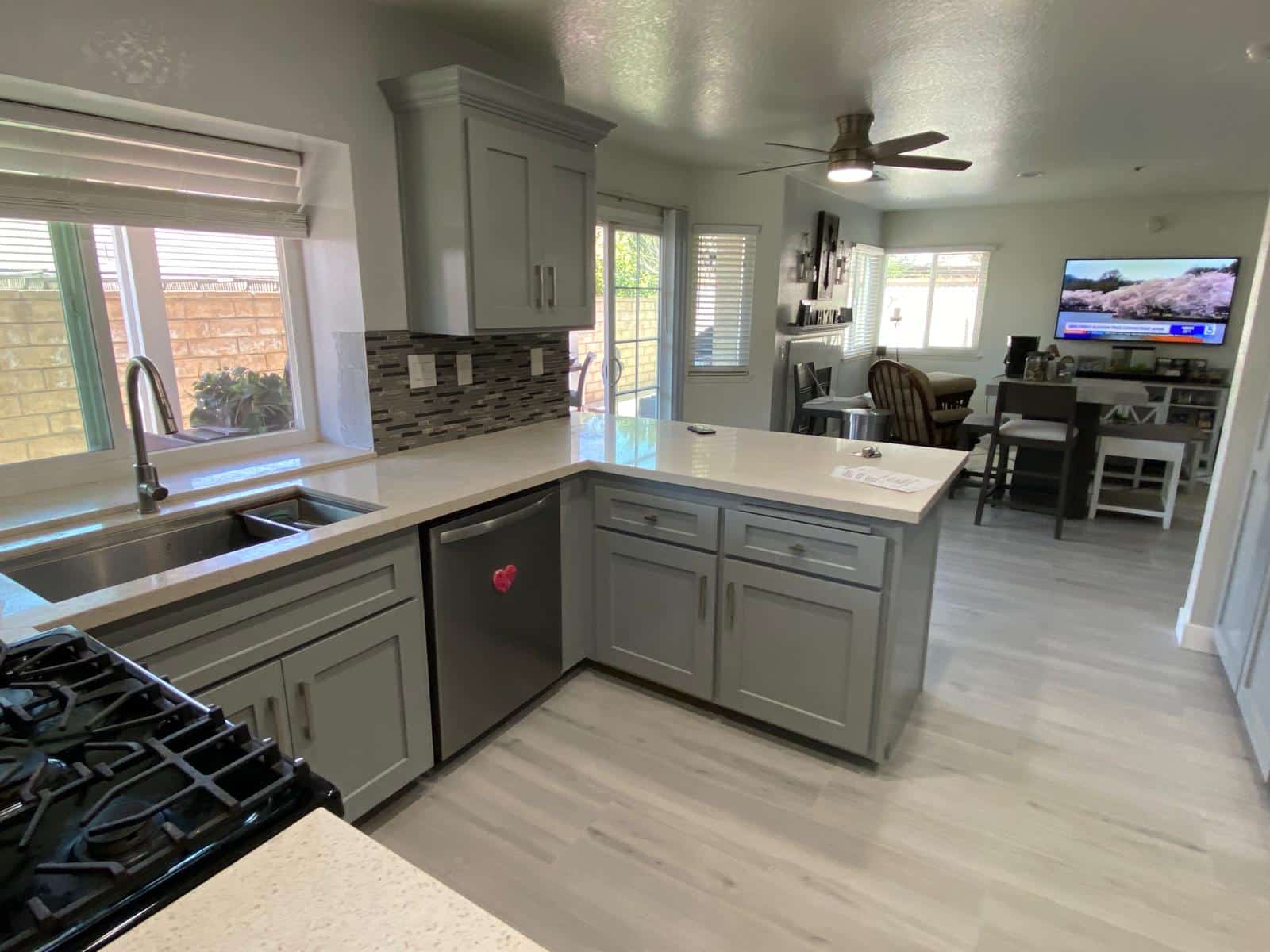 A modern kitchen and living area featuring gray shaker-style cabinetry, white quartz countertops, and stainless steel appliances including a gas range and dishwasher. The open-concept space flows into a family room with a flat-screen TV, and natural light streams through multiple windows and glass doors overlooking a brick patio. Light wood-look flooring and a bronze ceiling fan complete the contemporary renovation design.