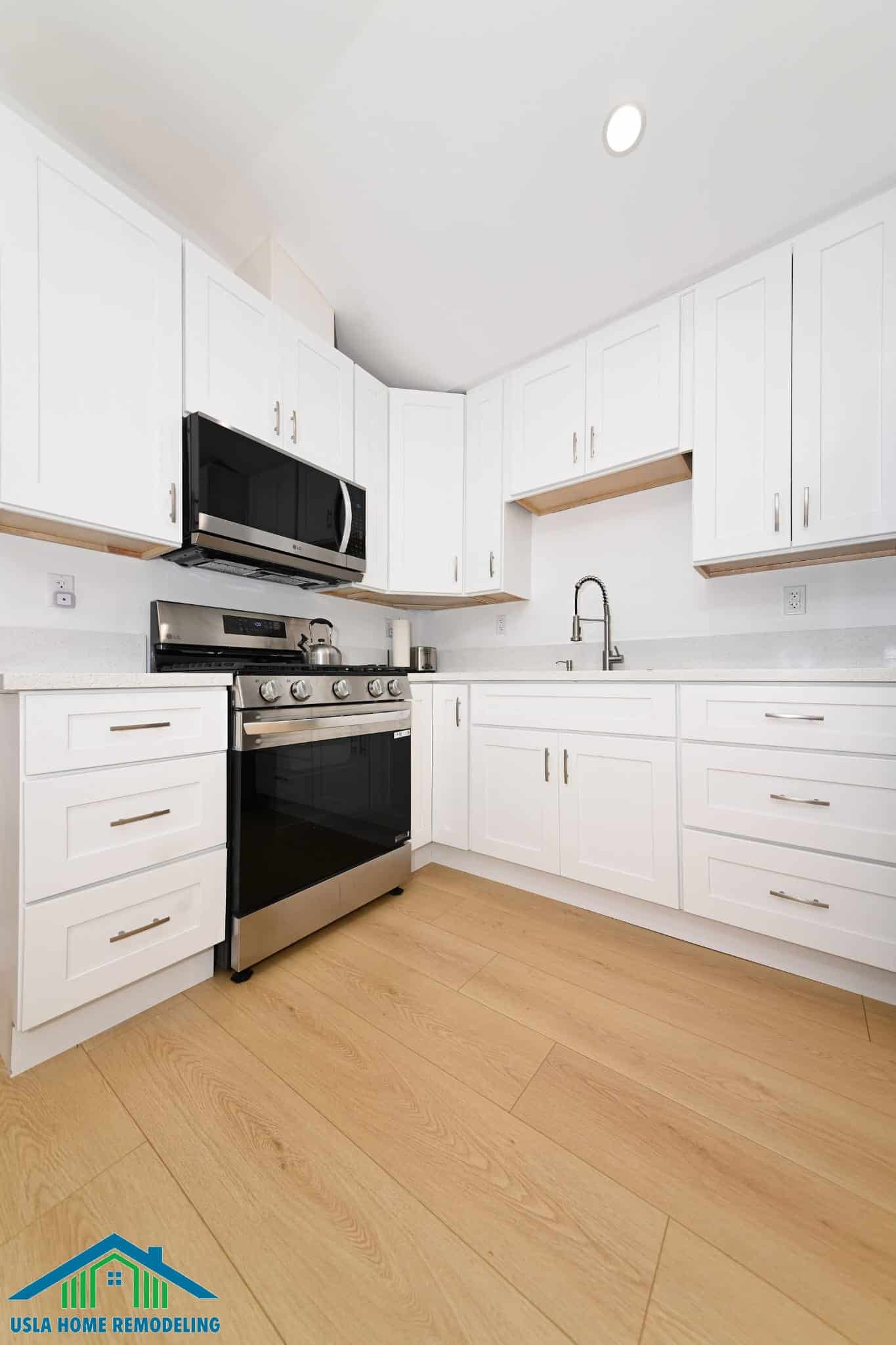 A modern kitchen features white shaker-style cabinets with natural wood accents and brushed nickel hardware, paired with stainless steel appliances including a black gas range with microwave above and a pull-down faucet at the sink. The bright space includes light wood-look flooring, white subway tile backsplash, and recessed ceiling lighting, showcasing a contemporary kitchen remodel. This design exemplifies professional home remodeling work with attention to detail and quality finishes.