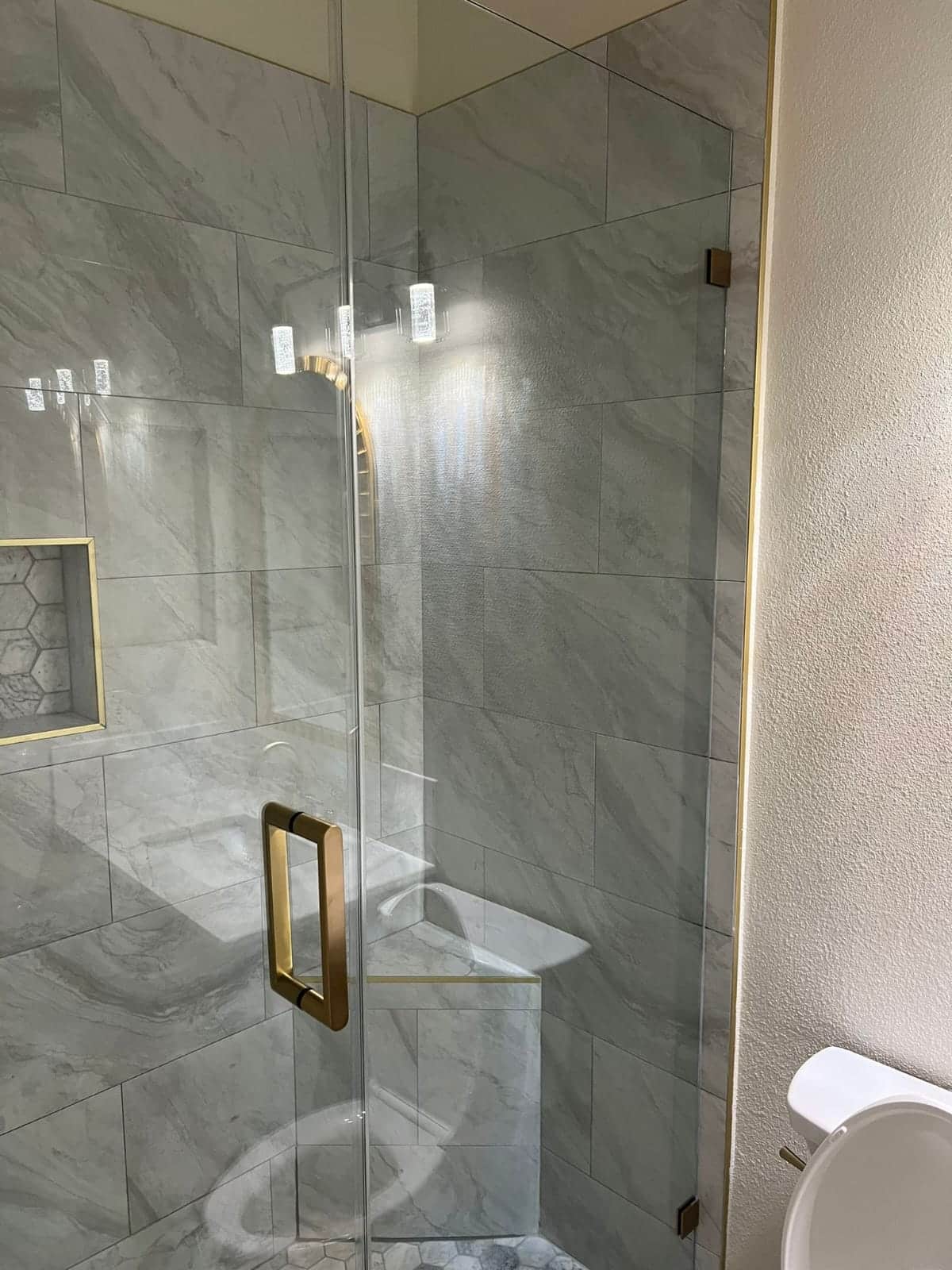 A modern bathroom shower enclosure featuring frameless glass doors with bronze hardware and a built-in bench seat. The shower walls are lined with large-format gray marble-look porcelain tiles arranged in a grid pattern, with recessed lighting fixtures mounted above. A white toilet is visible to the right of the enclosure, and the surrounding bathroom walls have a textured finish with brass-trimmed accents.