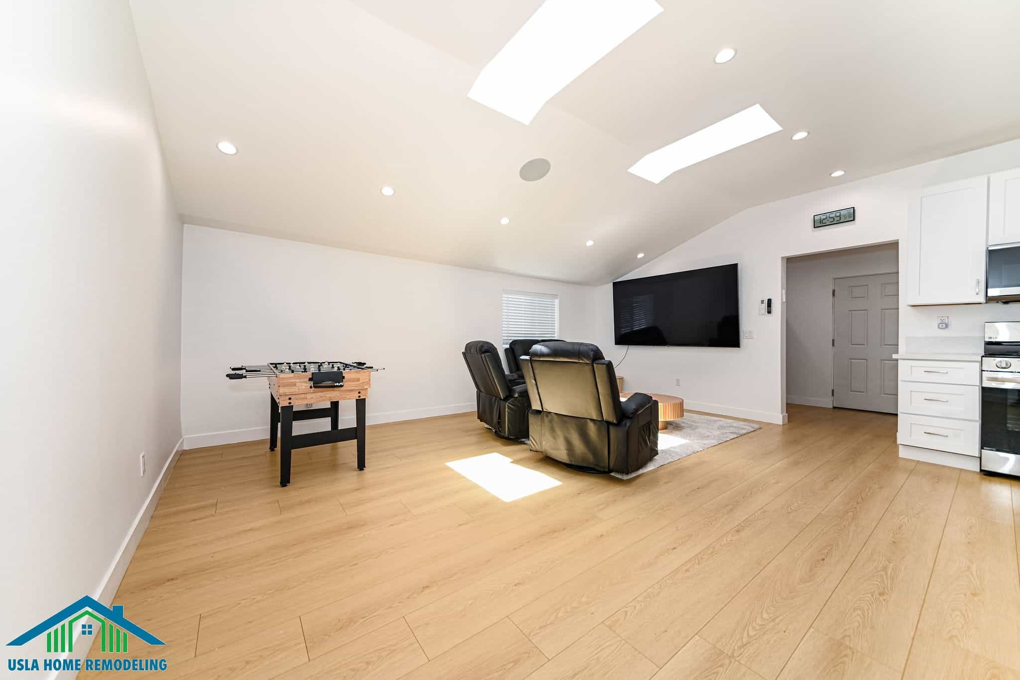 A modern, spacious basement recreation room features light wood flooring, recessed and skylight ceiling fixtures, and white walls with a wall-mounted flat-screen TV. The room is furnished with a black leather recliner, foosball table, and includes a kitchen area with white cabinetry and stainless steel appliances on the right side. This professionally remodeled space demonstrates a complete basement transformation suitable for entertainment and family gatherings.