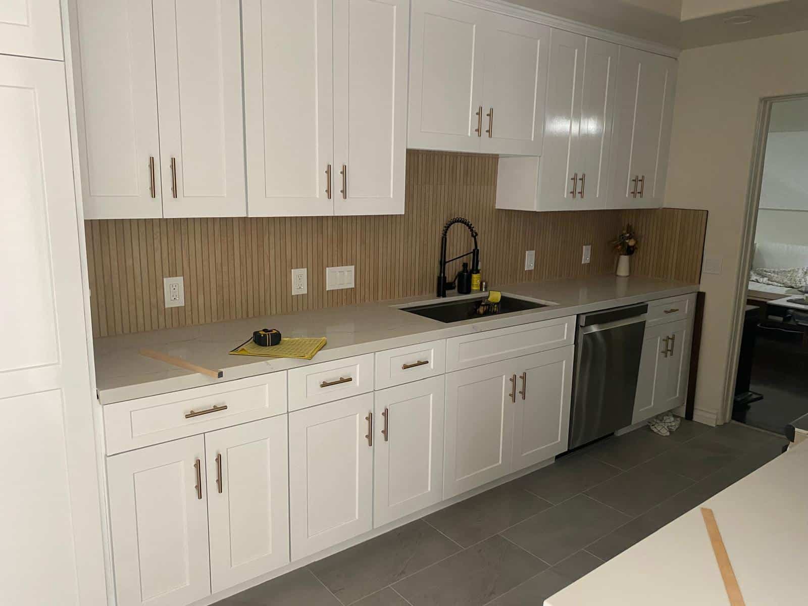 A modern kitchen remodel featuring white shaker-style cabinetry with brushed brass hardware, a warm tan vertical shiplap backsplash, and gray quartz countertops. The space includes stainless steel appliances including a built-in dishwasher and range, a black undermount sink with a matte black pull-down faucet, and dark tile flooring. The kitchen displays a clean contemporary design with ample storage and organized layout for both functionality and aesthetic appeal.