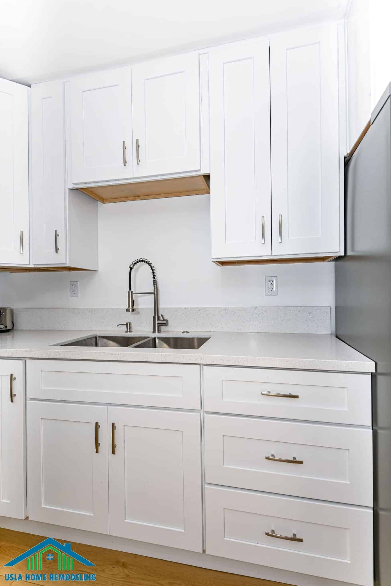 A modern kitchen featuring white shaker-style cabinetry with brass hardware, light gray quartz countertops, and a stainless steel double sink with a pull-down faucet. The kitchen includes upper cabinets with wood shelf accents, pendant lighting fixtures, and stainless steel appliances, showcasing a clean contemporary remodel design.