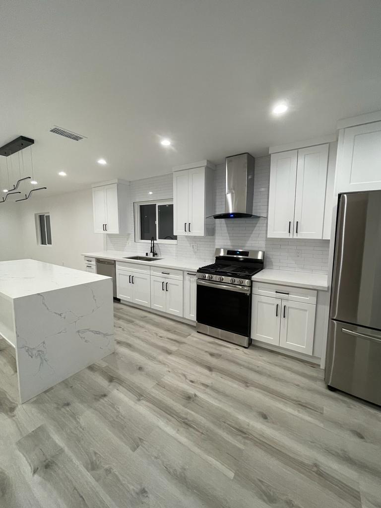 A modern kitchen remodel featuring white shaker-style cabinets, white subway tile backsplash, and stainless steel appliances including a black gas range and range hood. The kitchen includes a white waterfall-edge quartz island countertop and light wood-look vinyl plank flooring, with recessed lighting throughout the newly renovated space.