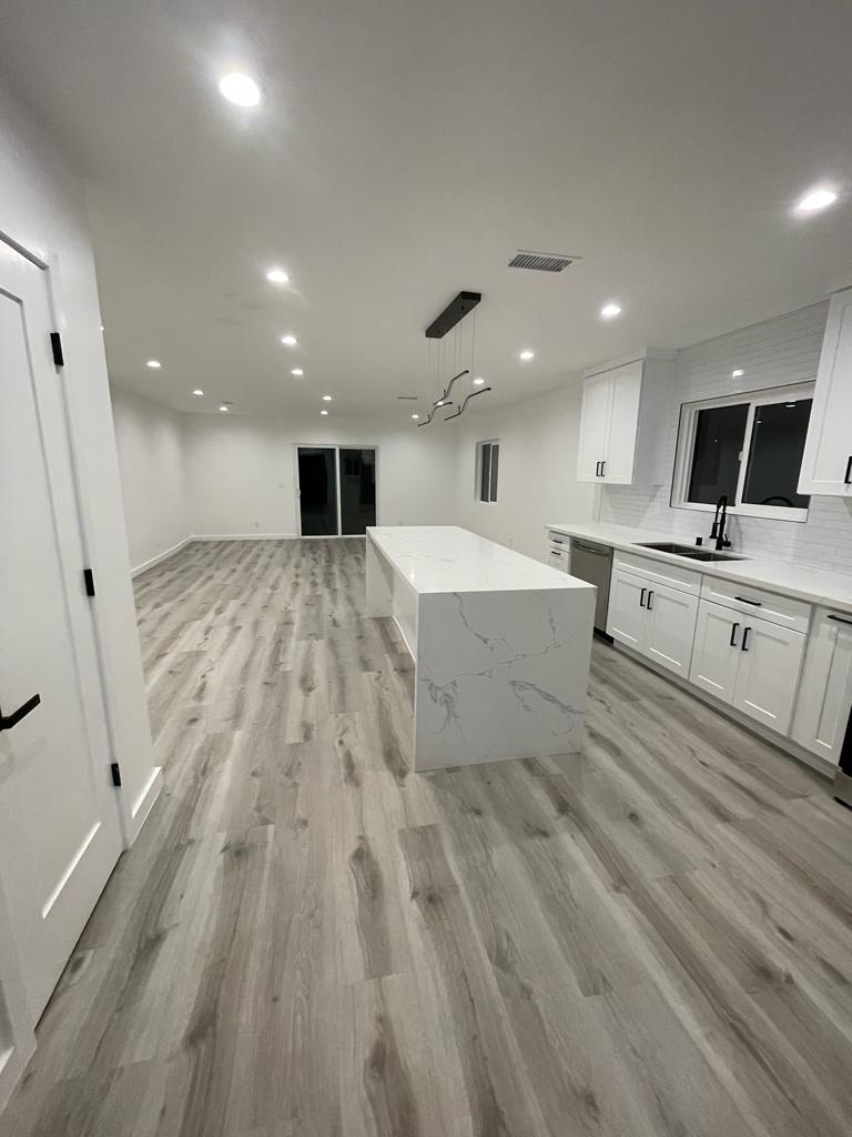 A modern, newly remodeled kitchen and living space featuring white cabinetry, a large white marble-look island, and gray wood-look vinyl plank flooring throughout. Recessed lighting is installed in the ceiling, and the open concept design connects to a spacious living area with glass doors visible in the background. White subway tile backsplash and stainless steel appliances complete the contemporary, clean aesthetic of this residential renovation project.