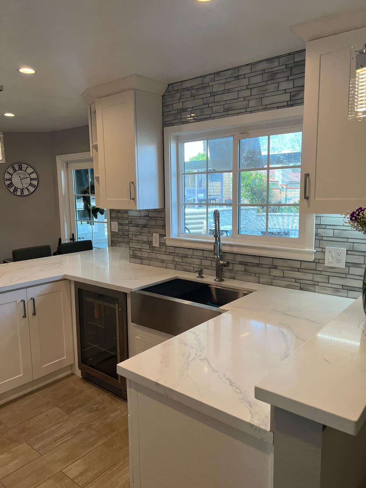 A modern kitchen remodel featuring white cabinetry, marble-look countertops, and a large island with seating. The backsplash displays gray subway tiles arranged in a staggered pattern, and a large window above the sink provides natural light and views of the exterior. Stainless steel appliances and fixtures complete the contemporary design.