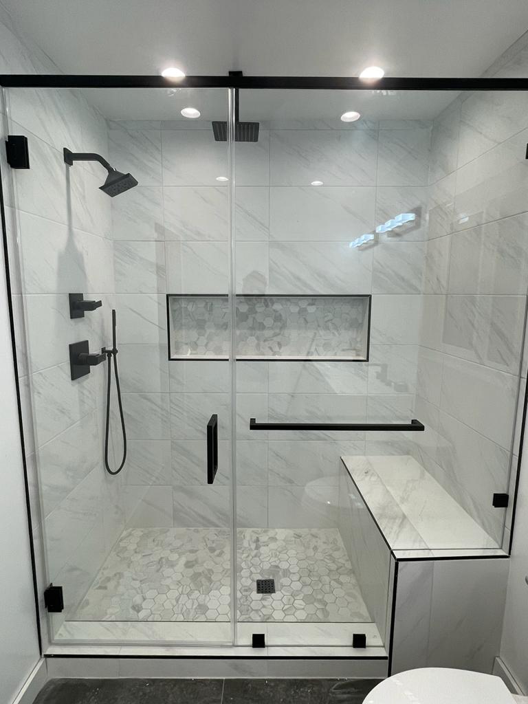 A modern bathroom shower enclosure featuring large white marble-look tiles on the walls, black framed glass doors, and a marble hexagonal mosaic tile floor. The shower is equipped with a black square showerhead, dual handle controls, a handheld sprayer, and includes a built-in bench seat and recessed shelving niche. Recessed lighting is installed in the ceiling and along the glass enclosure frame.