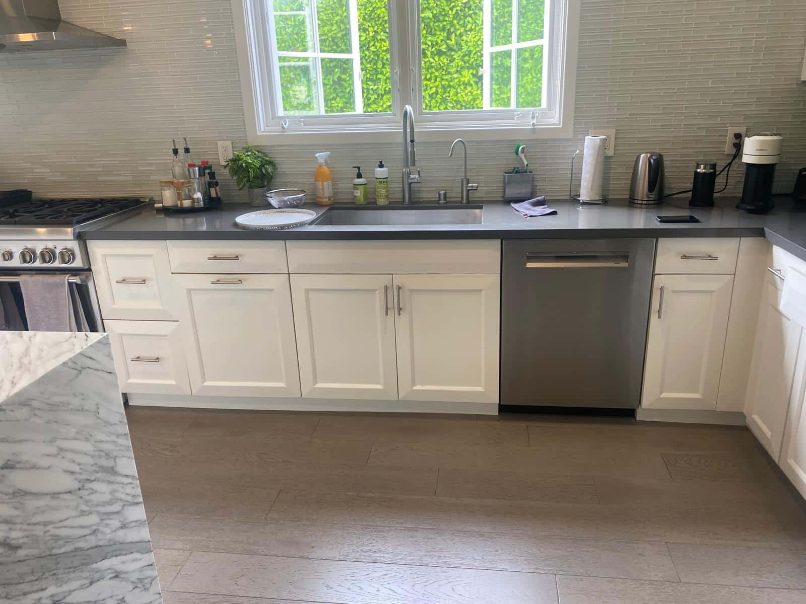 A modern kitchen featuring cream-colored cabinetry with stainless steel hardware and a gray quartz countertop, complete with a large sink and stainless steel dishwasher. The space includes light gray subway tile backsplash, professional gas range on the left, and abundant natural light from large windows overlooking green landscaping. Light wood-look tile flooring and marble countertops on the kitchen island complete the contemporary, clean aesthetic of this renovated kitchen.