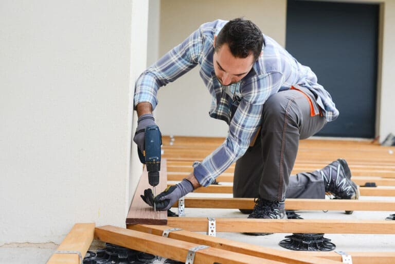 A man in a blue plaid shirt and work gloves uses a power drill to install hardwood flooring in an interior space. He is crouched over wooden planks, securing them to the subfloor while tools and materials are scattered around the work area. The setting appears to be a home renovation or remodeling project in progress.