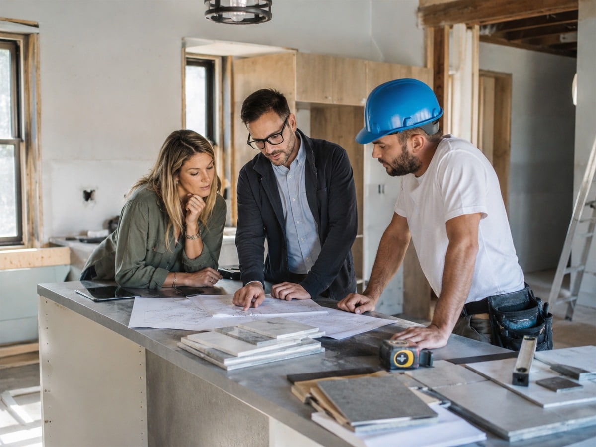Three professionals review construction blueprints and plans at a counter in an unfinished home interior. A woman in casual business attire and two men—one in professional clothing and one wearing a blue hard hat and tool belt—collaborate on the renovation project. The counter is scattered with architectural drawings, material samples, and construction tools in a bright, under-renovation space with exposed wooden framing.
