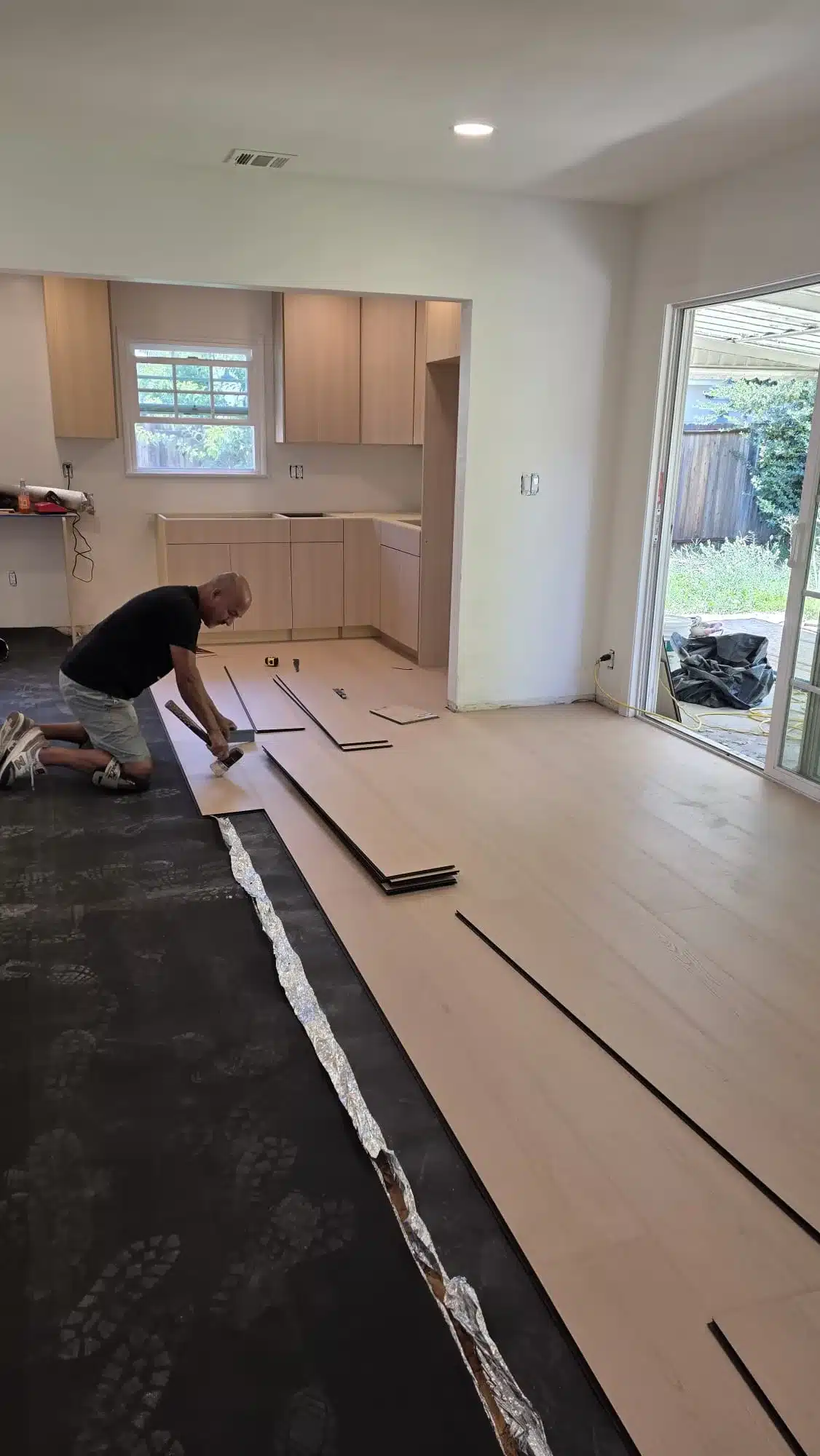 Worker installing light wood-tone plank flooring beside kitchen cabinets and a sliding glass patio door.