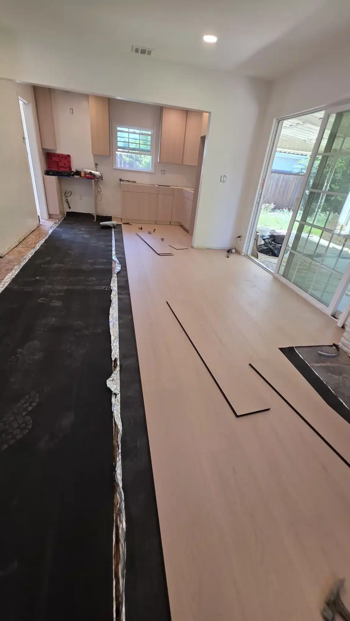 Partially installed light wood-tone plank flooring with exposed dark underlayment beside kitchen cabinets.