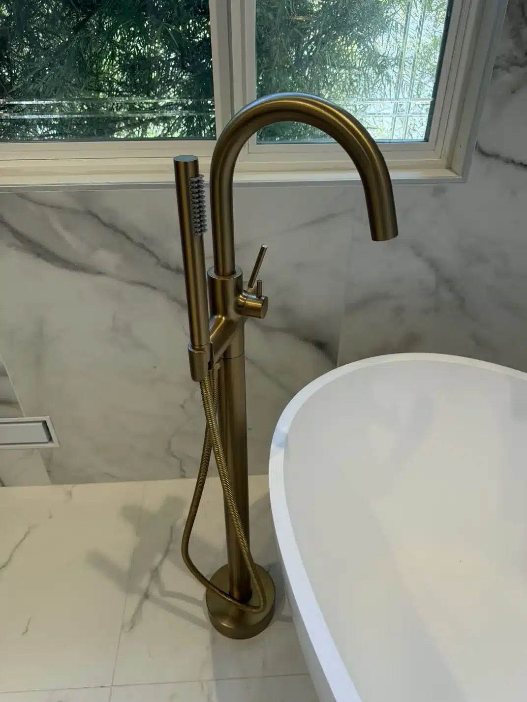 Brass-tone floor-mounted tub filler with handheld shower beside a white tub beneath a window.