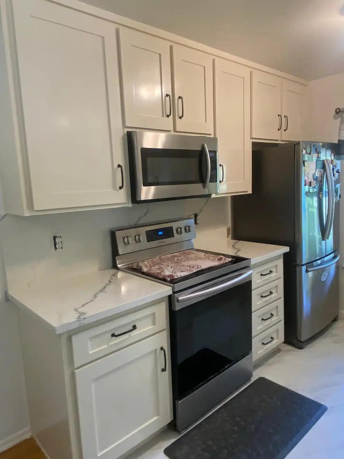 White shaker cabinets with black pulls, gray-veined counters, a stainless range and microwave, and uncovered outlets.