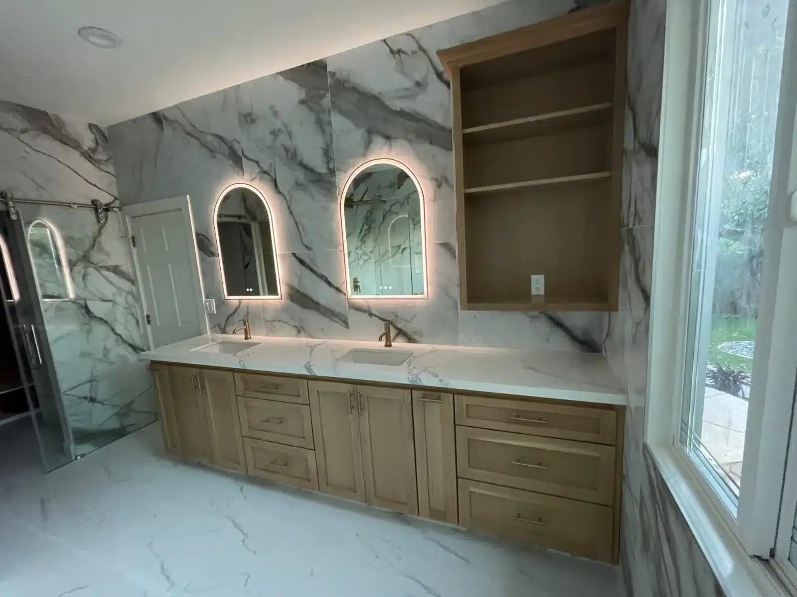 Bathroom with wooden dual-sink vanity, marble walls, and two illuminated arched mirrors
