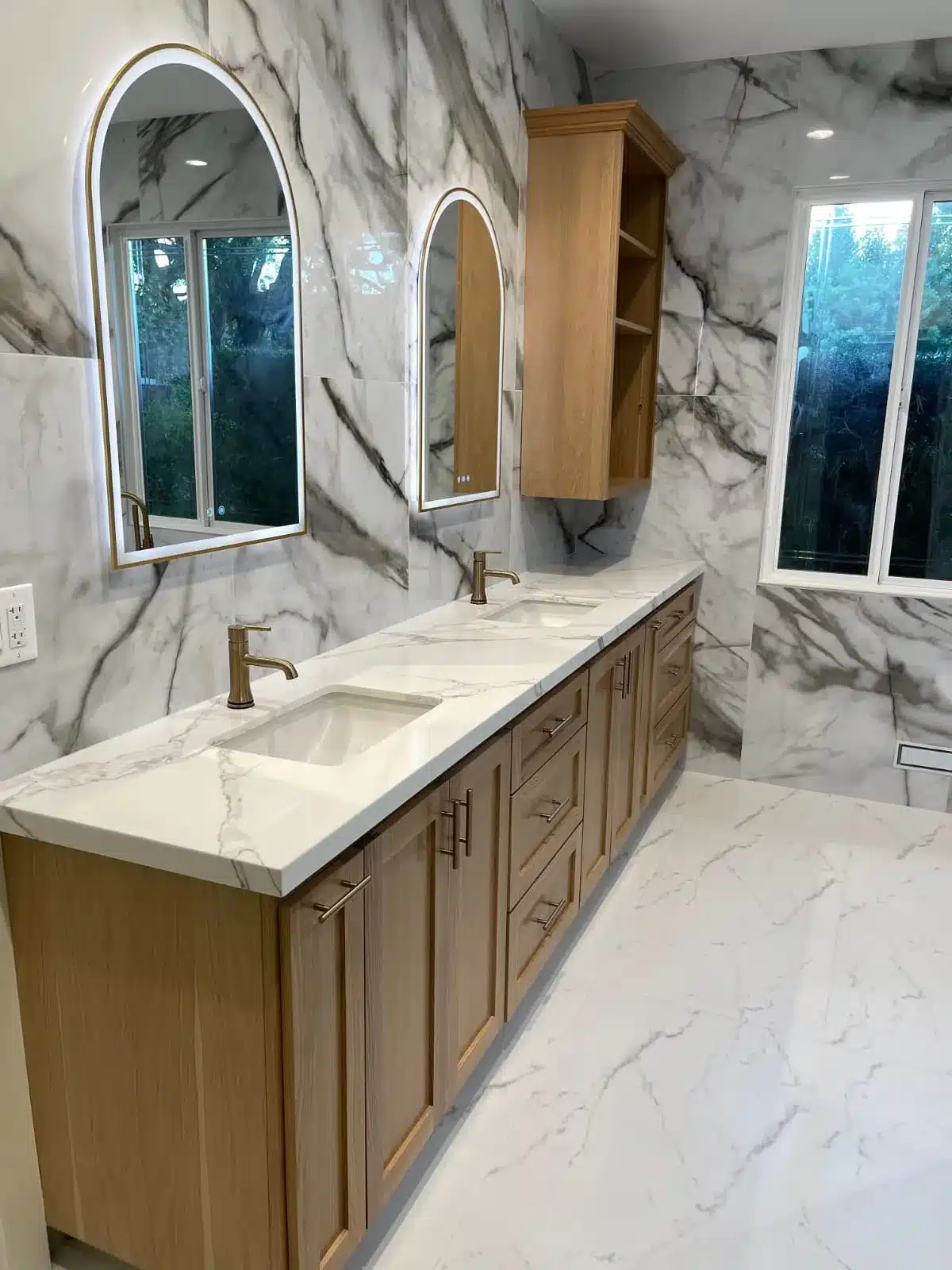 Bathroom with long wooden vanity, white marble counters, two arched illuminated mirrors, and marble tile walls